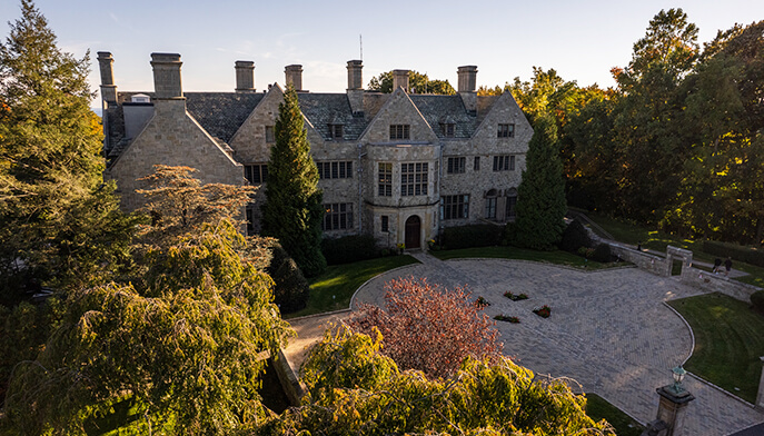 An aerial view of Bellarmine Hall, a large stone building with a gabled roof, surrounded by lush green trees and a parking lot with several cars on a sunny day.